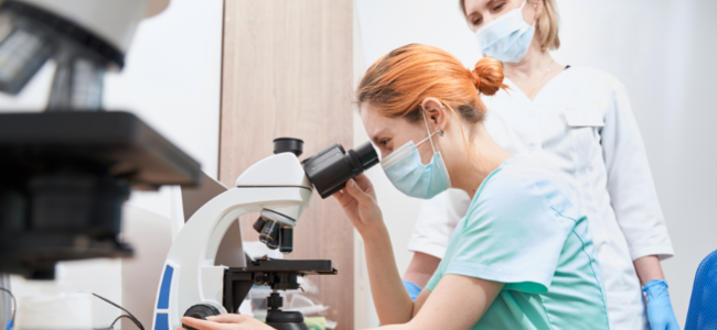 Una técnica observa a través de un microscopio en un laboratorio, asistida por otra persona, ambas usando mascarillas